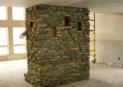 Field Stone - fireplace in middle of room small shelving build in with lots of windows in background and scaffolding