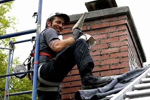 Tech repointing chimney smiling and has on safety gear ladder and scaffolding