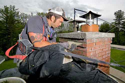 Tech with safety gear repointing chimney he has 2 tatoos on his arm and wearing a hat woods in background